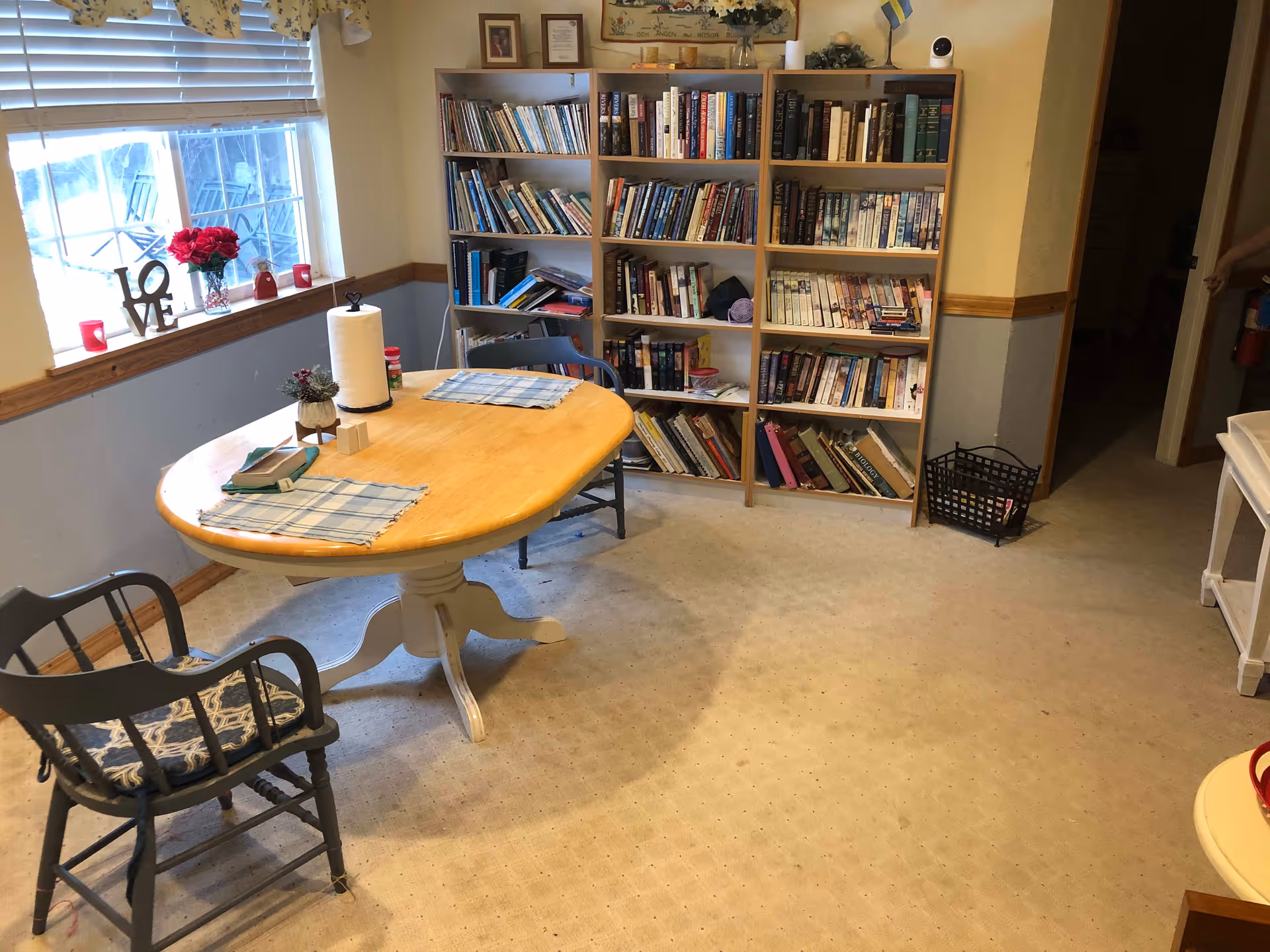 A cozy room with a wooden table and two chairs, a bookshelf filled with books against the wall, a window with blinds and floral curtains, and decorative items including a vase with red flowers and a LOVE sign on the windowsill.