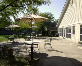 Outdoor patio area with several metal tables and chairs, each table shaded by large umbrellas. The patio is adjacent to a light-colored building with multiple windows and surrounded by trees and greenery.