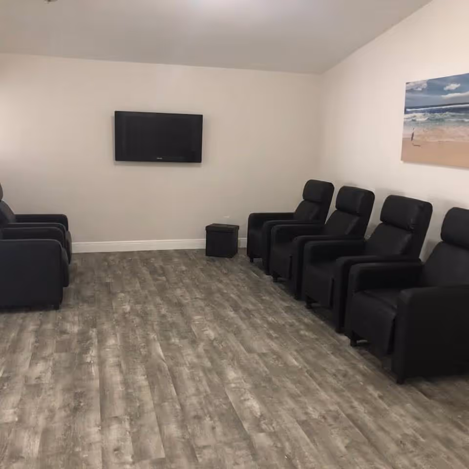 A common room with rows of black recliner chairs facing a wall-mounted TV and wood-look flooring.