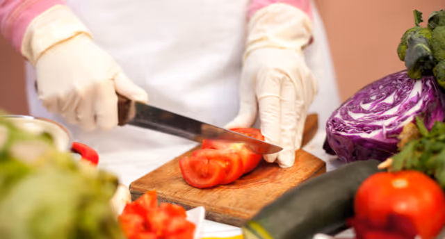 Gloved hands slice a red tomato on a wooden cutting board in a kitchen with other vegetables nearby.