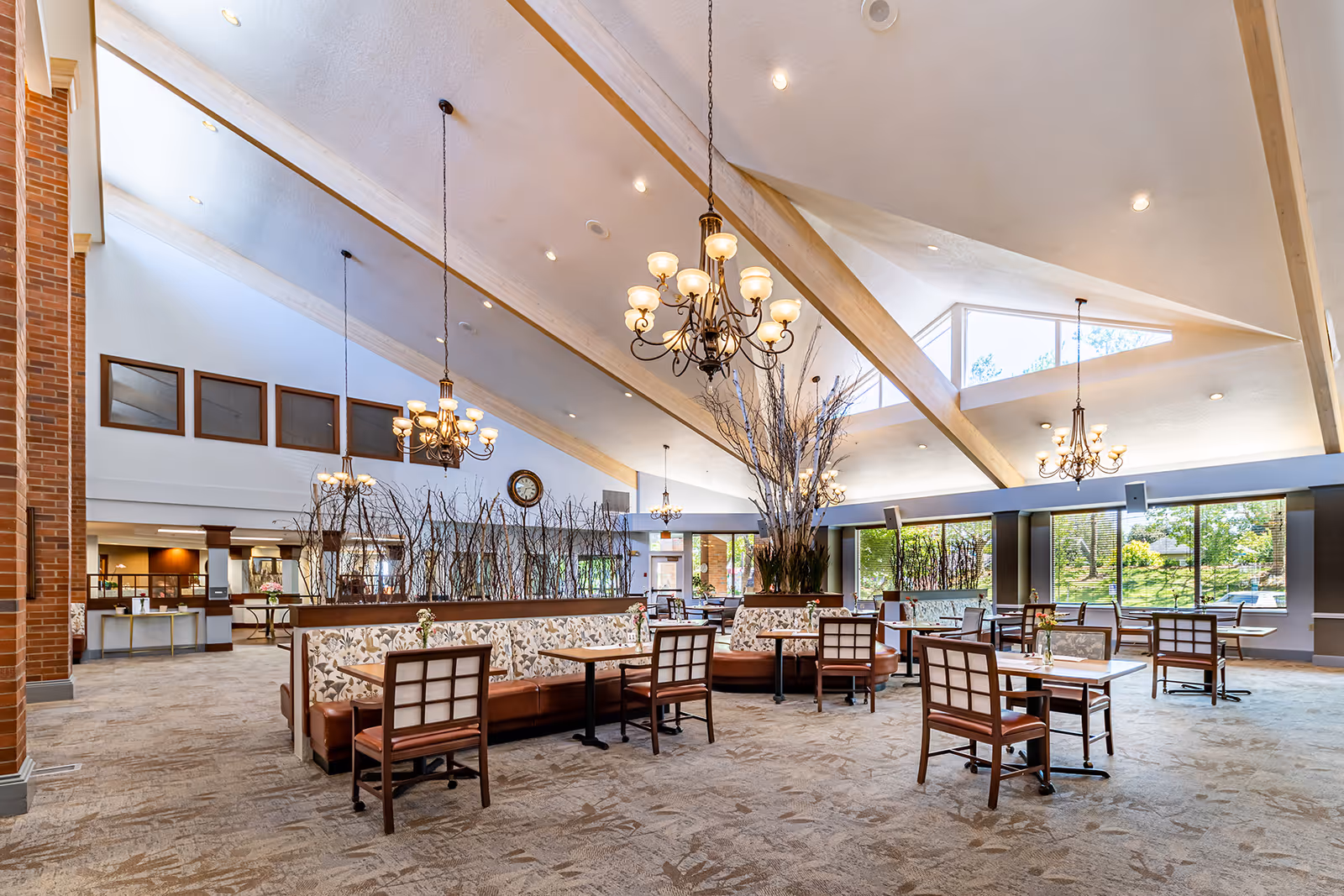 Spacious dining area with high vaulted ceilings featuring wooden beams and multiple chandeliers. The room has large windows allowing natural light to brighten the space, with tables and chairs arranged neatly around cushioned booth seating decorated with floral patterns. There are decorative branches placed in planters on top of the booth dividers, and a clock is mounted on the far wall.