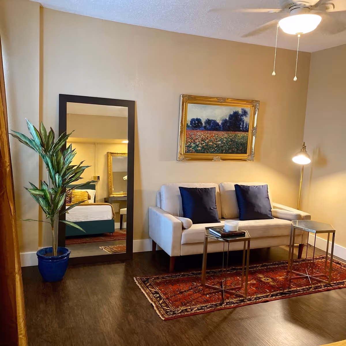 A cozy living room area with a beige loveseat adorned with two dark blue pillows, a red patterned rug underneath, and two small metal side tables in front. A tall floor lamp with a white shade stands to the right, and a large rectangular mirror with a dark frame leans against the left wall, reflecting a bed and dresser. A potted green plant in a blue pot is placed next to the mirror. Above the loveseat hangs a framed painting of a field with trees in the background. The room has wooden flooring and beige walls with a ceiling fan overhead.