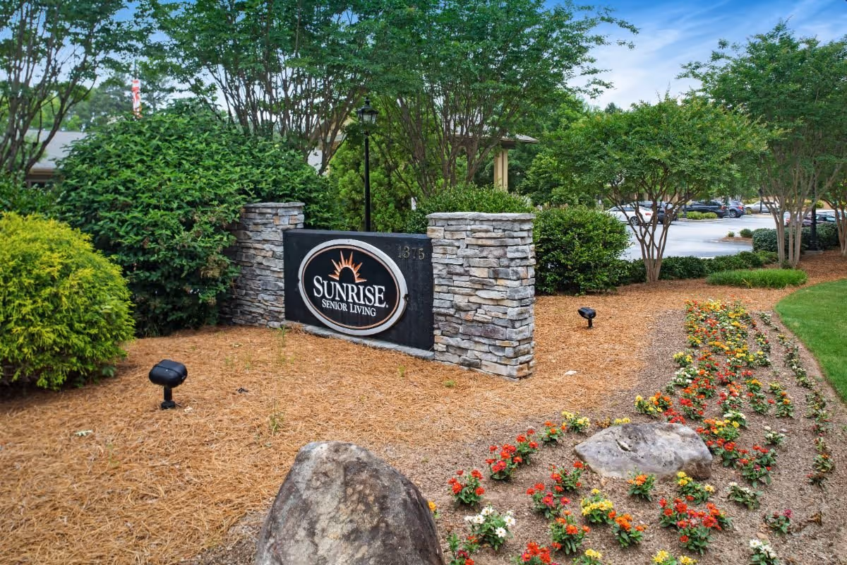 Stone entrance sign that reads "Sunrise Senior Living" amid landscaped beds, shrubs, and a nearby parking area.