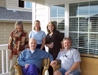 Five adults posing together on a patio outside a residential building. Three women are standing behind two seated men, all smiling and casually dressed. The background shows windows with white blinds and part of the building exterior.