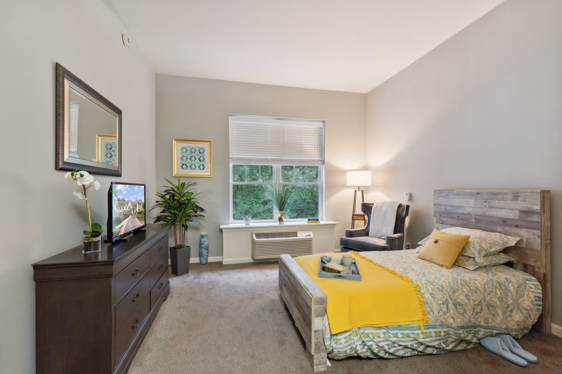 A cozy bedroom in an assisted living facility featuring a wooden bed with patterned bedding and a yellow throw blanket. A dark wooden dresser with a mirror above it is on the left side, decorated with a potted orchid and a TV. A window with blinds lets in natural light, and a comfortable armchair with a floor lamp is positioned in the corner. The room has neutral-colored walls and carpeted flooring.