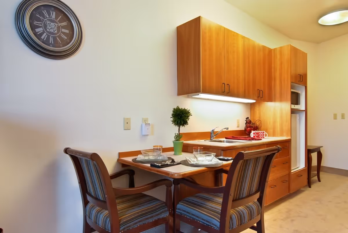 A small kitchen area with wooden cabinets, a sink, and a countertop. In front of the countertop is a dining table set for two with striped cushioned chairs. A small potted plant is on the table. A wall clock with Roman numerals hangs on the wall above the table.