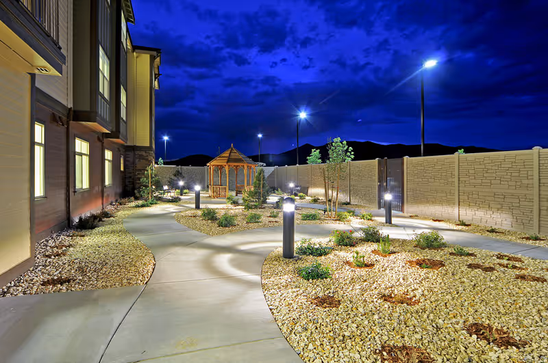 Well-lit outdoor courtyard at night with a winding concrete path, lamp posts, landscaped rock beds, a wooden gazebo, and the side of a building.