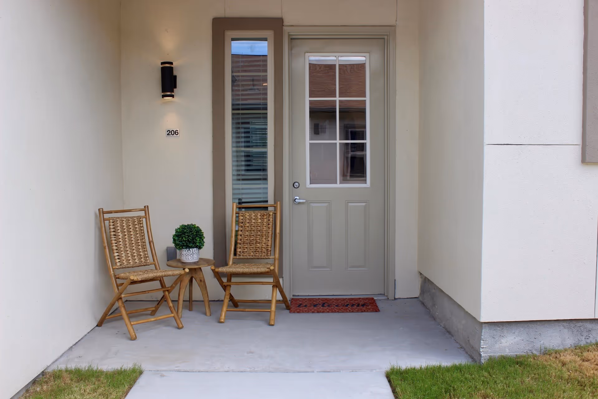 A small porch area outside an apartment or room with a beige door featuring a window with multiple panes. Two woven wooden chairs and a small round wooden table with a potted plant are placed on the porch. The wall beside the door has a light fixture and the number 206. There is a welcome mat in front of the door and grass on either side of the concrete walkway leading to the porch.