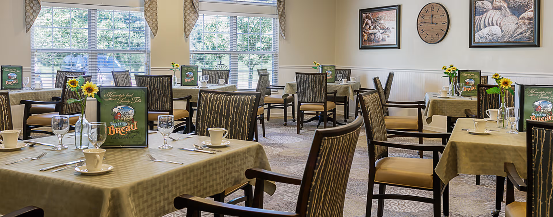 A bright dining room with several tables covered in beige tablecloths, each set with cups, glasses, silverware, and small vases holding sunflowers. The room has large windows with patterned valances letting in natural light, and framed artwork and a clock on the walls.
