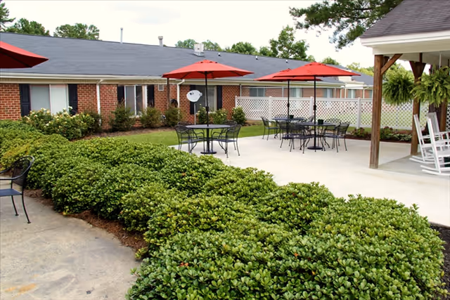 Outdoor patio area at Durham Ridge Assisted Living with several black metal tables and chairs under red umbrellas, surrounded by green bushes and a brick building in the background. There is a covered porch area with white rocking chairs and hanging ferns.