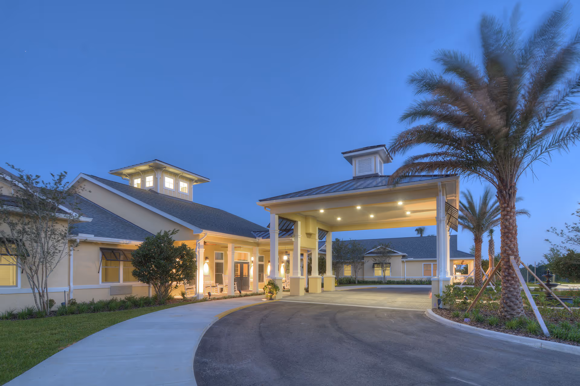 Exterior view of Benton House of Clermont senior living facility at dusk, showing a covered entrance with columns, illuminated windows, palm trees, and a curved driveway.