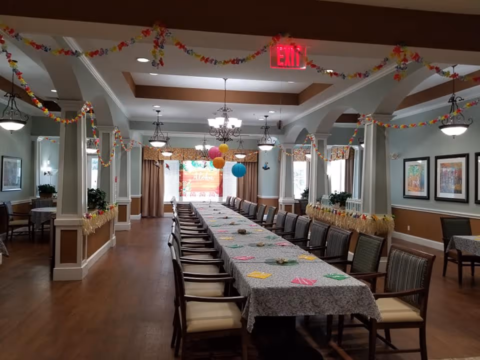 A long dining table covered with a patterned tablecloth and surrounded by chairs in a decorated dining room. The room features wooden floors, columns, chandeliers, and hanging colorful paper garlands and lanterns. There are framed pictures on the walls and windows with curtains at the far end.