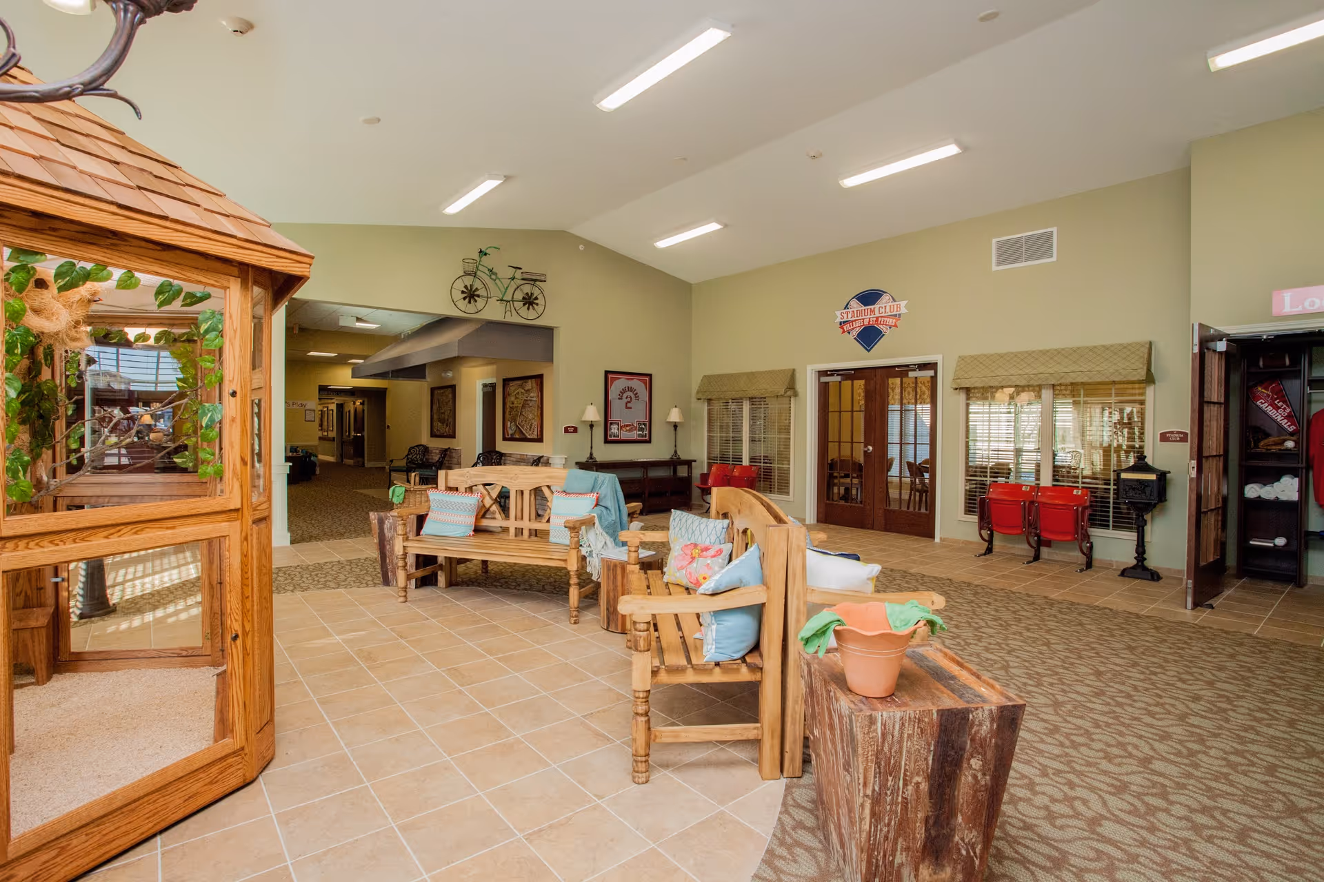 A spacious and well-lit common area in a senior living facility featuring wooden benches with colorful cushions, a wooden gazebo structure with plants inside, and decorative elements such as framed pictures and a vintage bicycle wall art. The room has tiled and carpeted flooring, beige walls, and large windows with blinds allowing natural light to enter.