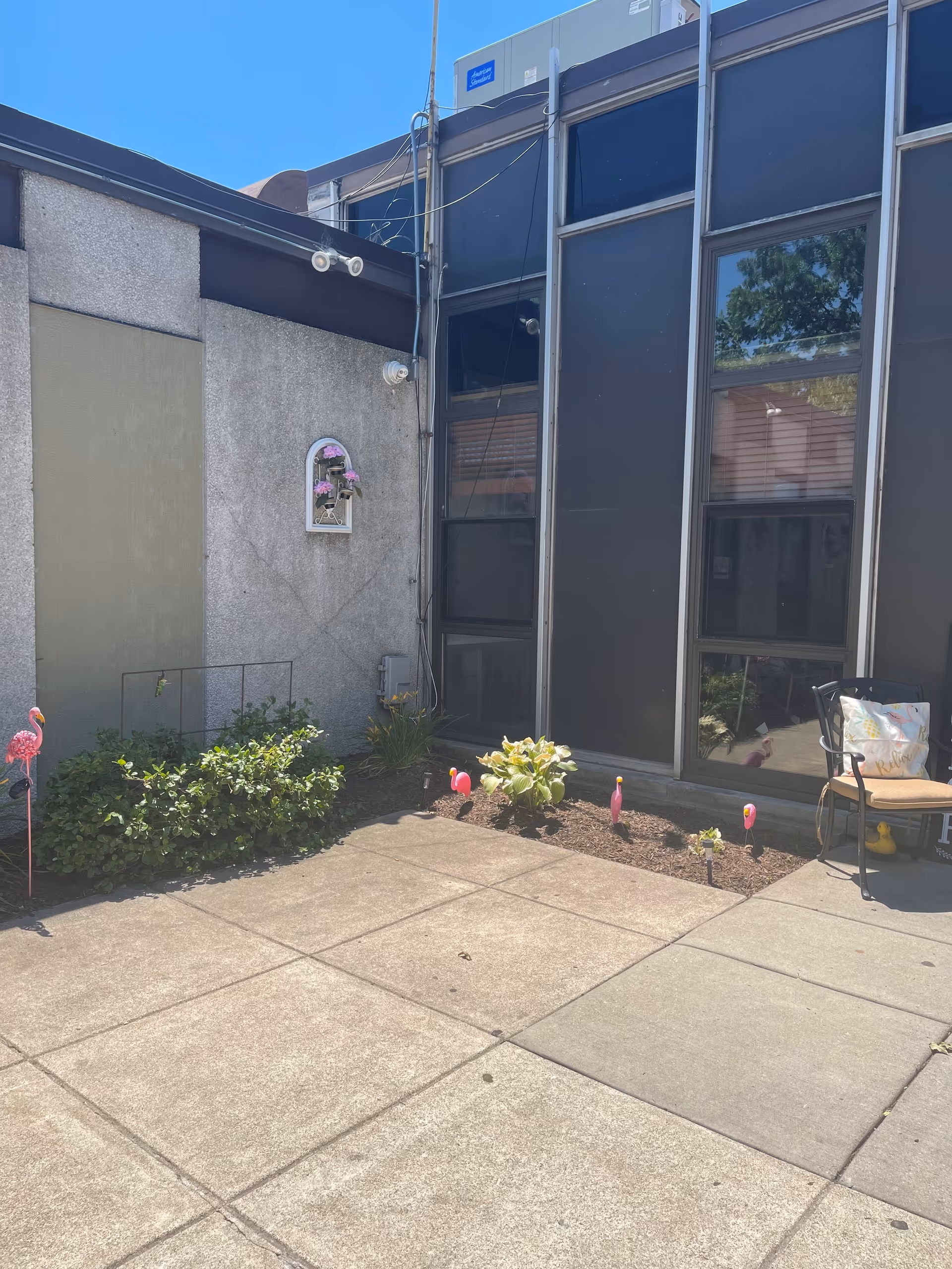 Outdoor patio area with concrete tiles, small garden beds with plants and pink flamingo decorations, a chair with a cushion, and a building with large windows in the background.