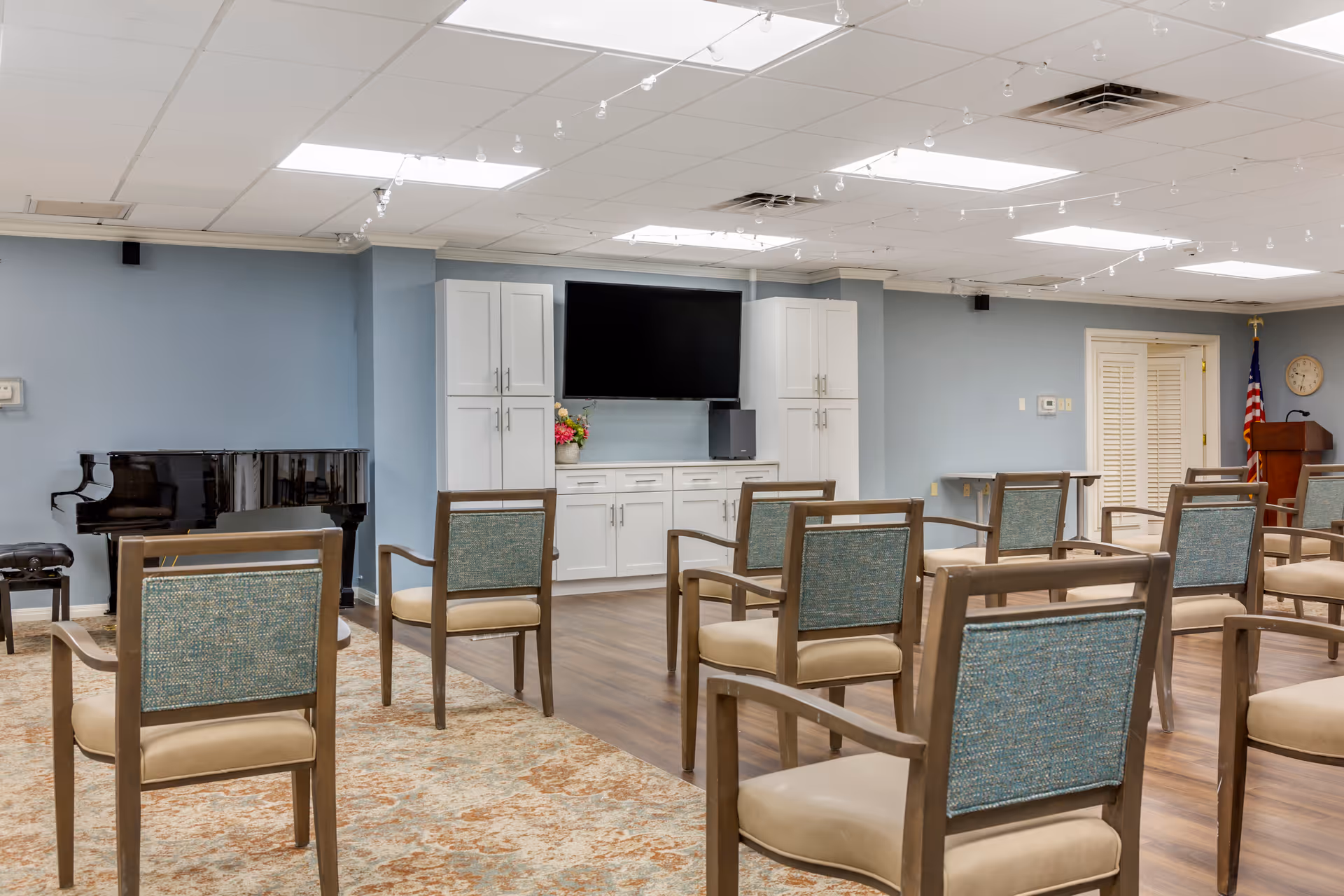 A bright multipurpose activity room with rows of chairs facing a wall-mounted TV, white cabinetry, and a black piano.