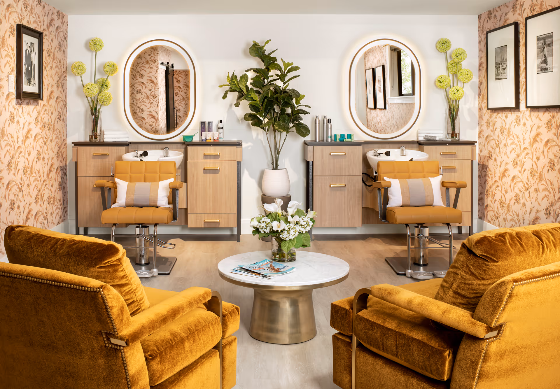 Bright salon-style interior with two styling chairs and wash basins, round mirrors, mustard armchairs and a central coffee table.