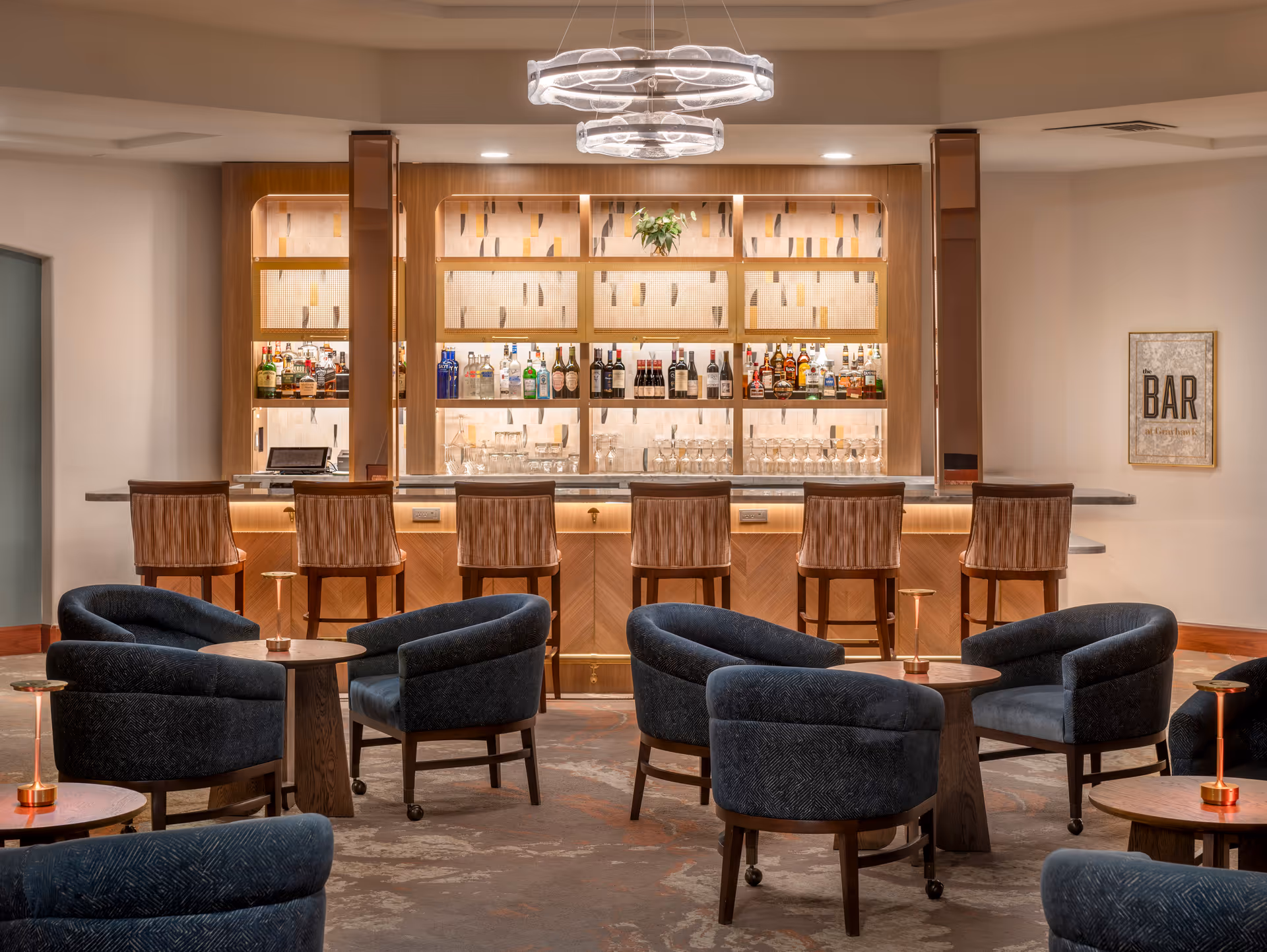 A modern bar area with a wooden counter and six high chairs. Behind the bar, shelves display various bottles of liquor and glassware, illuminated with soft lighting. In front of the bar, there are several round wooden tables surrounded by dark blue upholstered chairs. A contemporary circular chandelier hangs from the ceiling, and a framed sign on the wall reads 'BAR'.