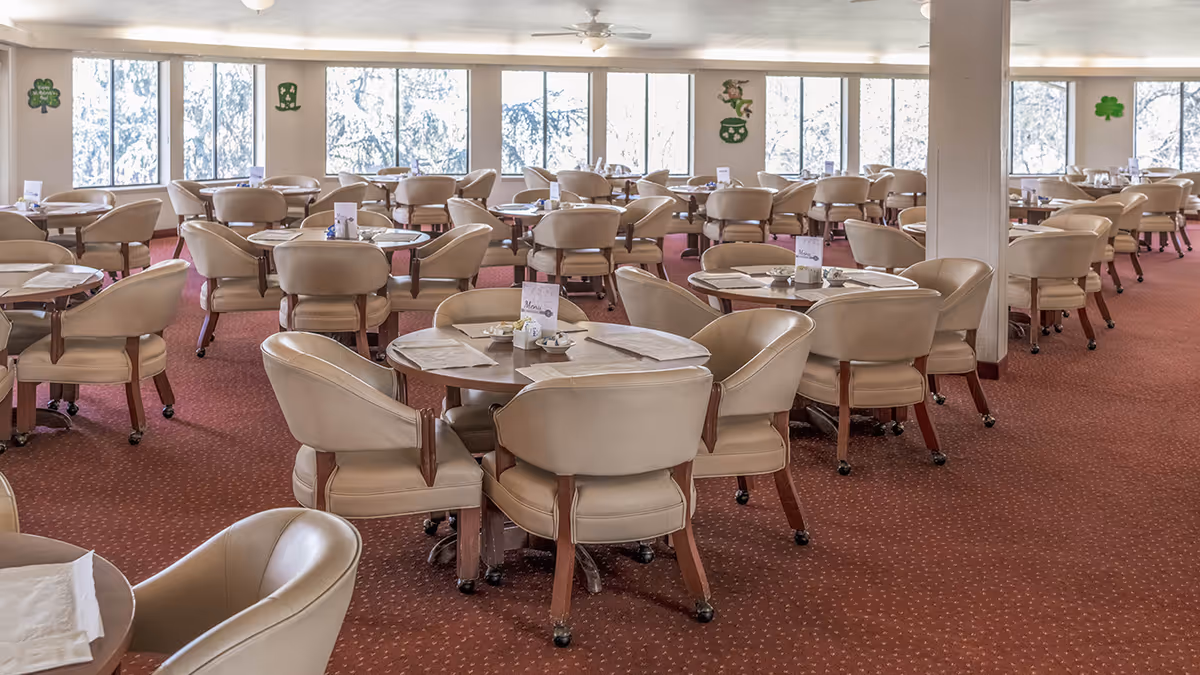 A spacious dining room with multiple round tables surrounded by beige cushioned chairs on wheels. Each table has menus, napkins, and small condiment containers. Large windows line the back wall, letting in natural light. The walls are decorated with St. Patrick's Day themed decorations including shamrocks and leprechaun hats. The carpet is a reddish-brown color with a subtle pattern.