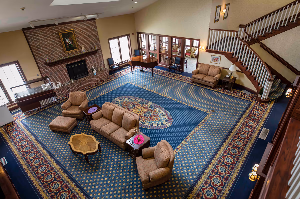 Spacious communal living room with patterned carpet, sofas and chairs around a coffee table, a brick fireplace, grand piano, and a staircase.