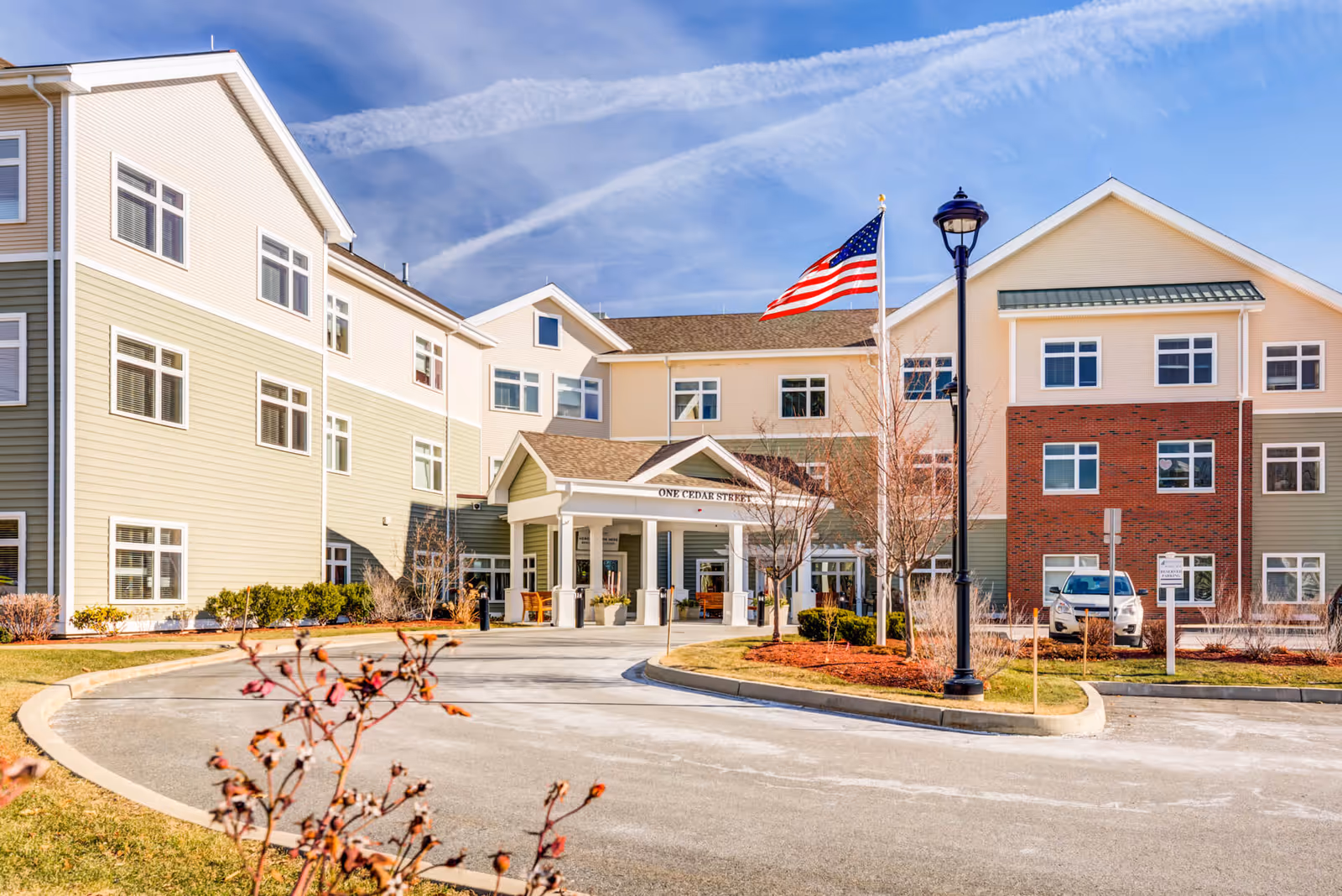 Front exterior view of a senior living facility building with beige and brick facade, multiple windows, an American flag on a flagpole, a street lamp, and a curved driveway leading to the entrance under a covered porch.