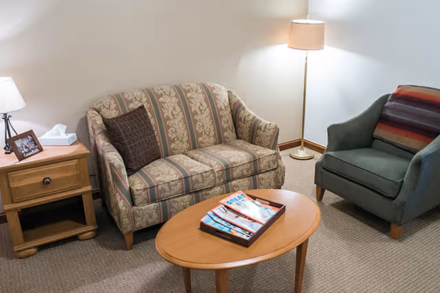 A cozy living room area with a patterned loveseat, a green armchair with a striped blanket draped over it, a wooden oval coffee table with magazines on a tray, a wooden side table with a lamp, tissue box, and framed photo, and a tall floor lamp in the corner.