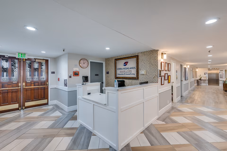 Reception area inside Serra Highlands Senior Living facility featuring a white reception desk, a wall-mounted sign with the facility name and logo, wooden double doors with glass panels, a wall clock, and a long hallway with framed pictures and an elevator at the end.