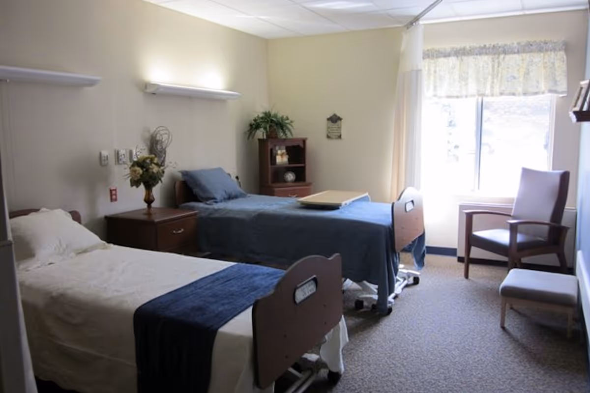 Two-bed senior resident room with adjustable beds, a nightstand, chair, window and simple decor.