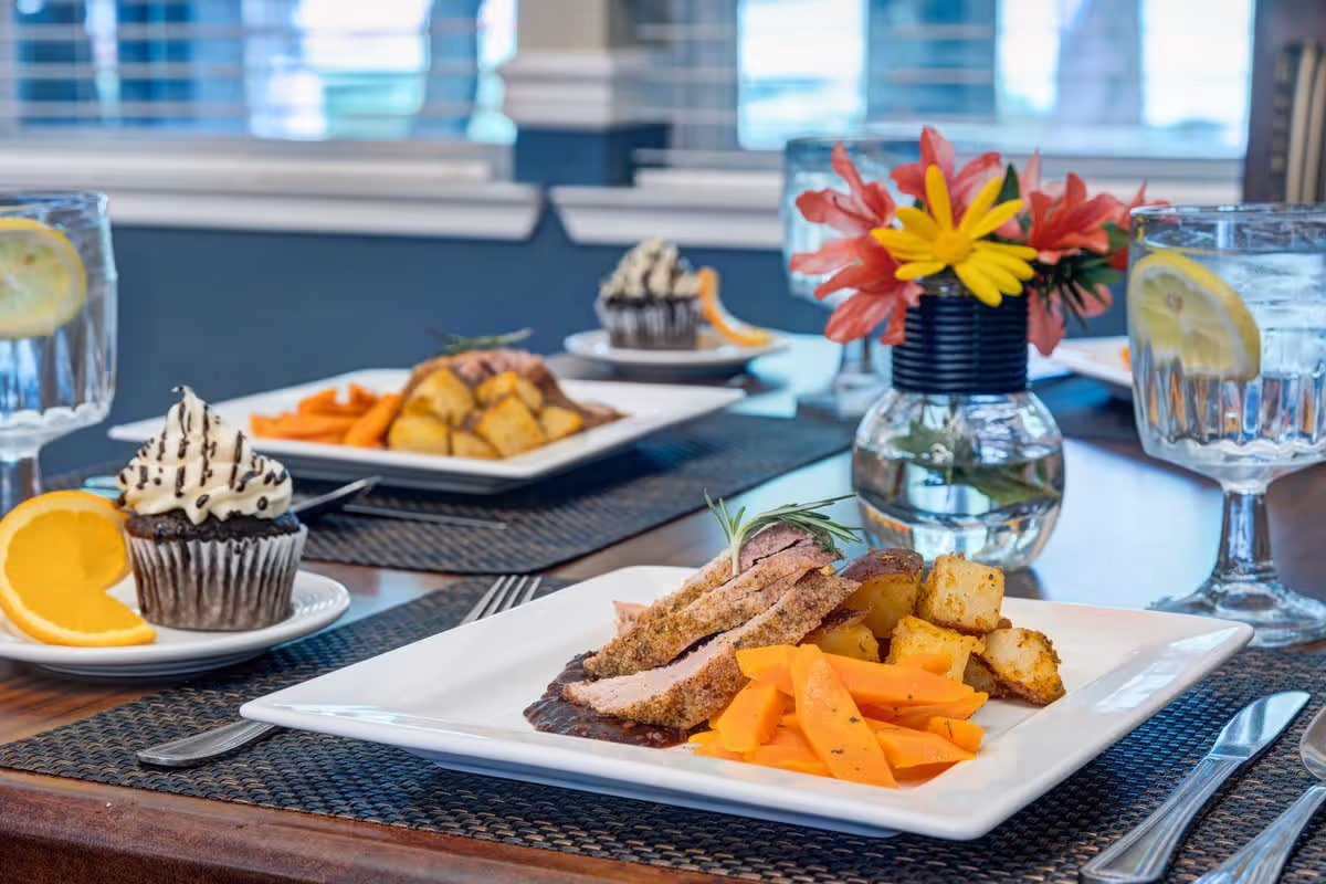 A dining table set with plates of food including sliced meat, roasted potatoes, and cooked carrots. Each place setting has a glass of water with a lemon slice and a chocolate cupcake with whipped cream and chocolate drizzle, garnished with an orange slice. A small vase with red and yellow flowers is placed in the center of the table.
