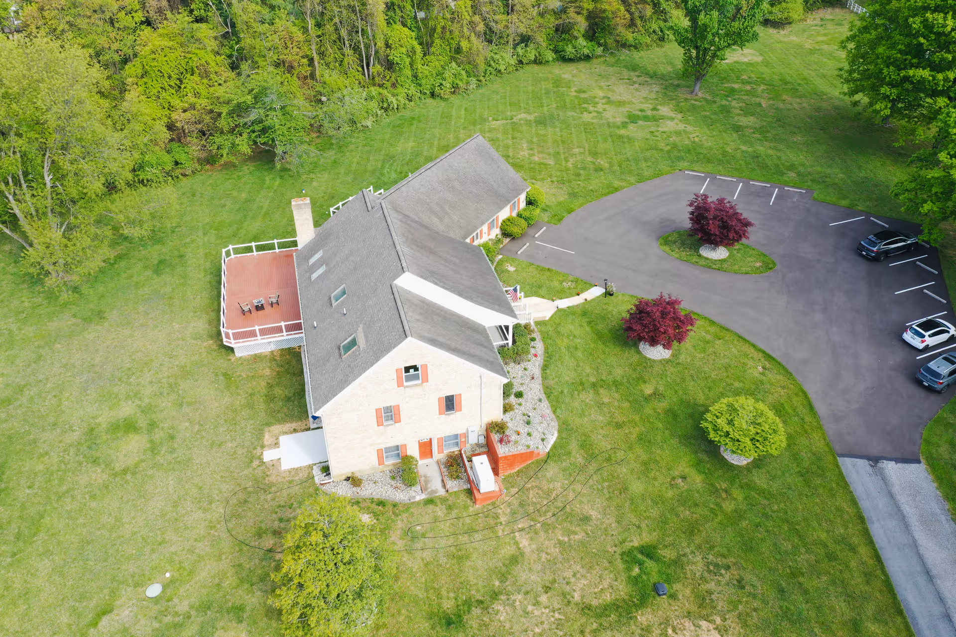 Aerial view of a large two-story residential building surrounded by green grass and trees. The building has a gray roof, light-colored brick walls, and red shutters. There is a wooden deck with outdoor furniture on one side and a paved parking area with several cars and landscaped islands with small trees nearby.