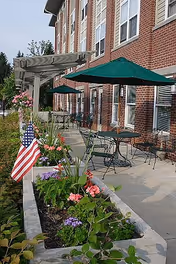 Outdoor patio along a brick retirement residence with tables and green umbrellas, flower planters and an American flag.