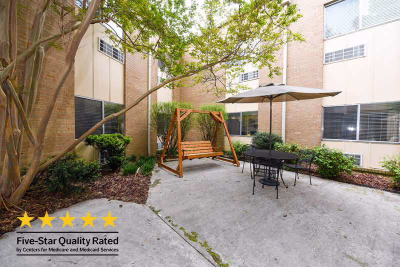 Outdoor courtyard area with a wooden swing, a round metal table with four chairs, and a large umbrella. The space is surrounded by brick walls with windows and some greenery including bushes and a tree.