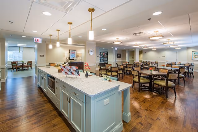 A spacious dining area in a senior living facility with multiple wooden tables and chairs arranged neatly on a polished wooden floor. In the foreground, there is a large kitchen island with a granite countertop, pendant lights hanging above, and built-in appliances. The room is well-lit with ceiling lights and has light-colored walls with some framed artwork. An exit sign is visible in the background near a doorway leading to another room with additional seating.