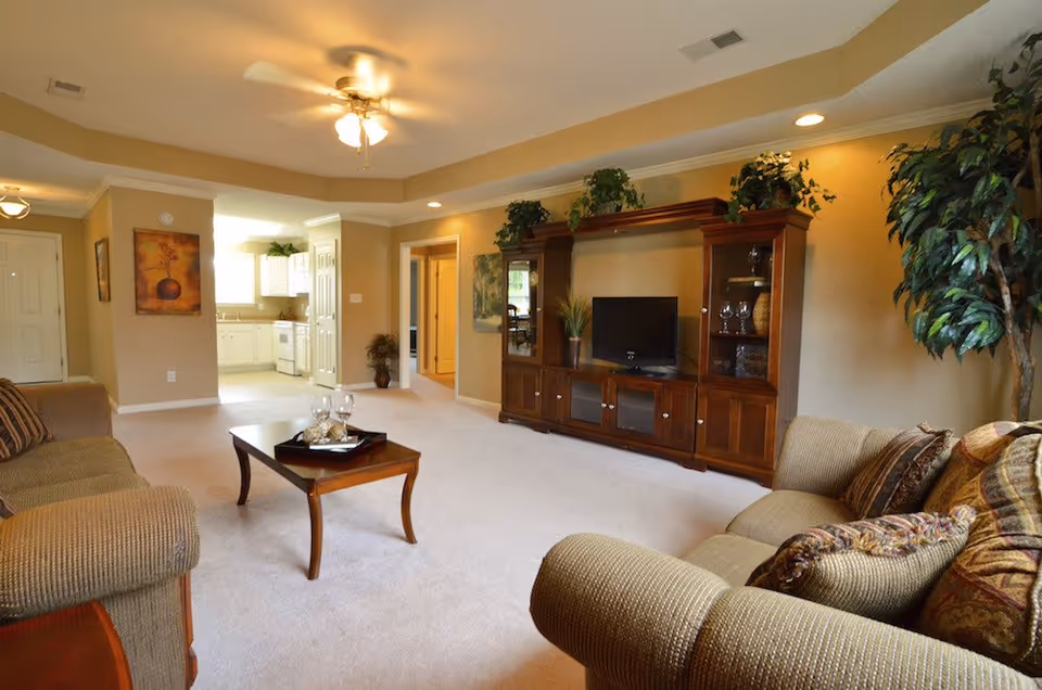 A cozy living room with beige walls and carpet, featuring two cushioned sofas with patterned pillows, a wooden coffee table with a tray and glasses, a large wooden entertainment center with a TV and decorative plants, and a ceiling fan with lights. The room opens into a bright kitchen area with white cabinets.
