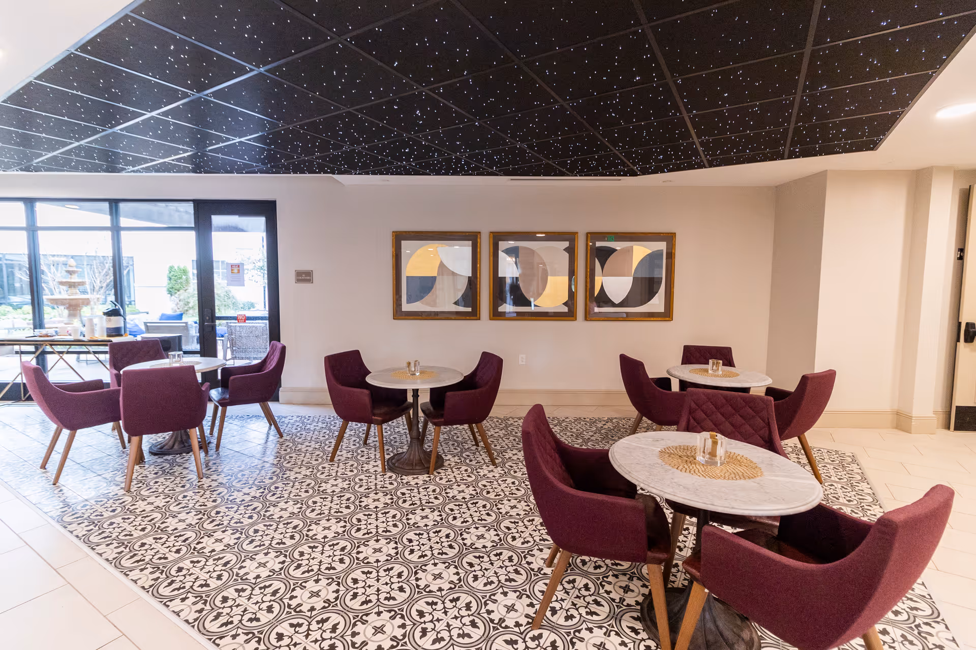 A modern indoor seating area with round marble tables and purple upholstered chairs arranged on a patterned tile floor. The ceiling above the seating area has a black design with small white dots resembling stars. Three abstract framed artworks hang on the wall, and large glass doors lead to an outdoor patio with additional seating and a water fountain.
