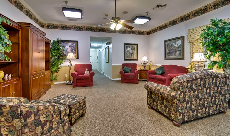 A cozy senior living facility common area with patterned armchairs and ottomans, red upholstered armchairs and sofa, wooden cabinets, framed artwork on the walls, potted plants, and table lamps providing warm lighting. A ceiling fan with lights is mounted above, and a hallway is visible in the background.