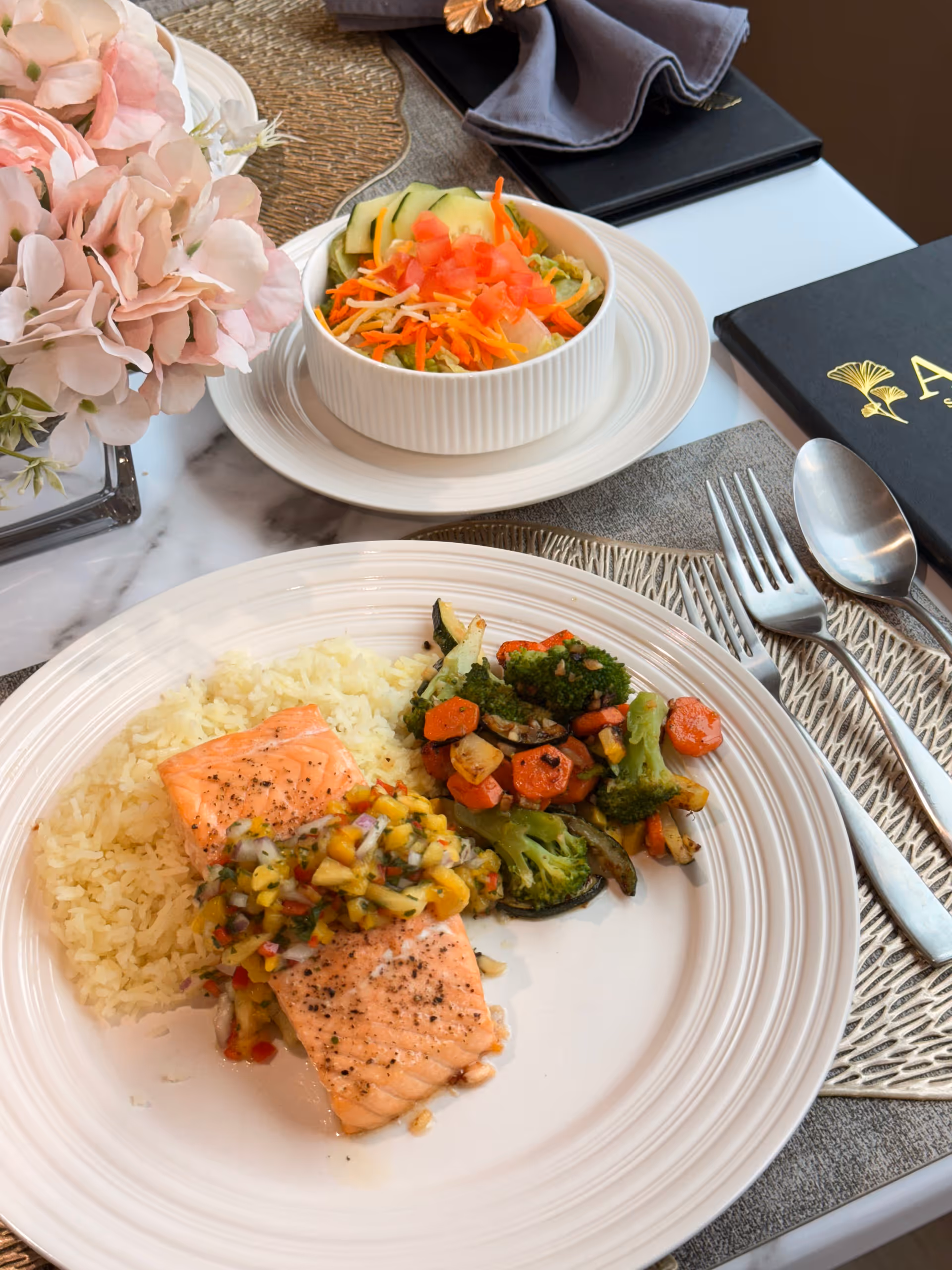 Plated salmon with rice and roasted vegetables plus a small salad and floral centerpiece on a set dining table.