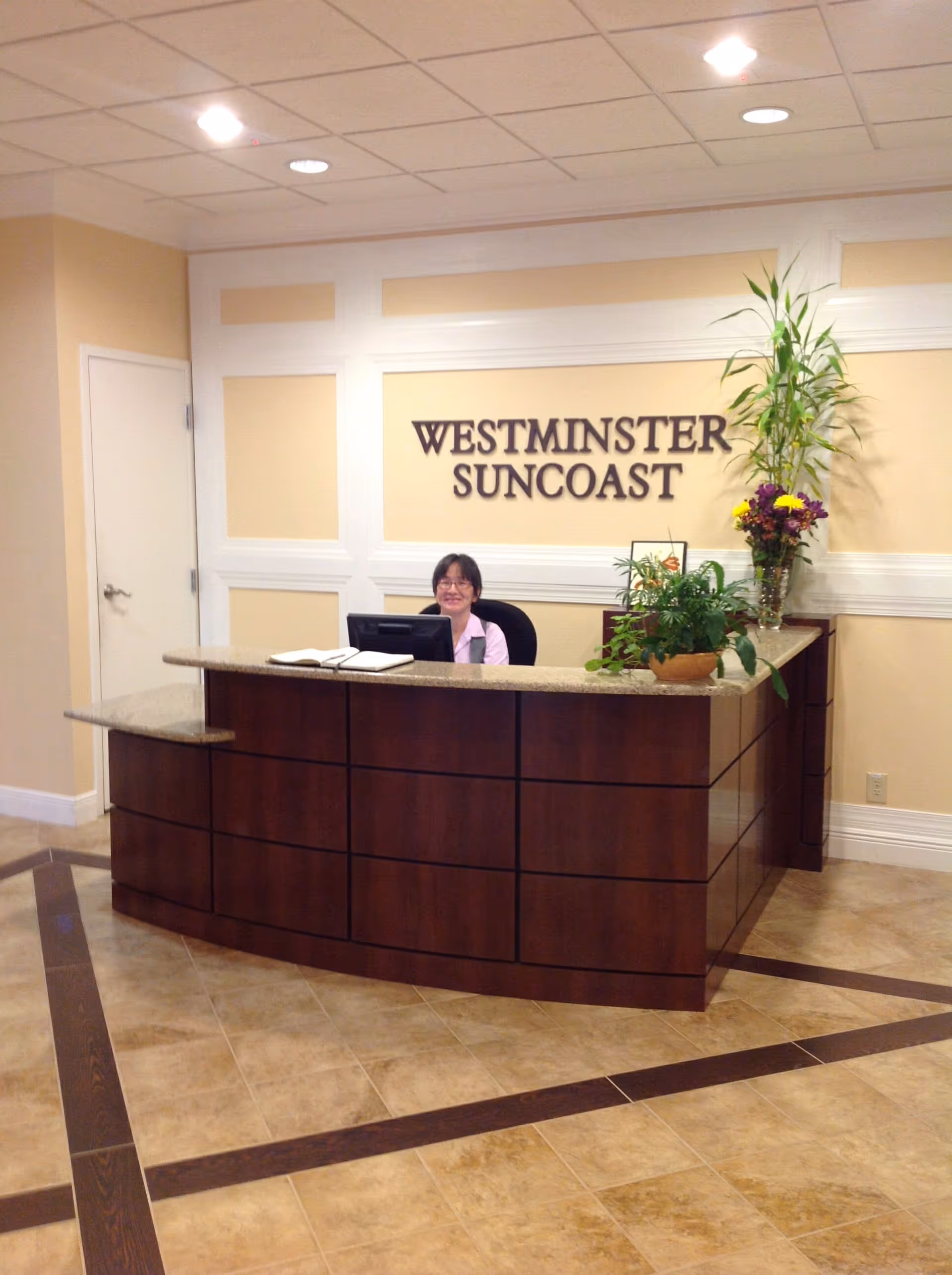 Reception area of Westminster Suncoast with a smiling receptionist sitting behind a wooden desk. The wall behind the desk has the facility name displayed, and there are plants on the desk and in the corner.