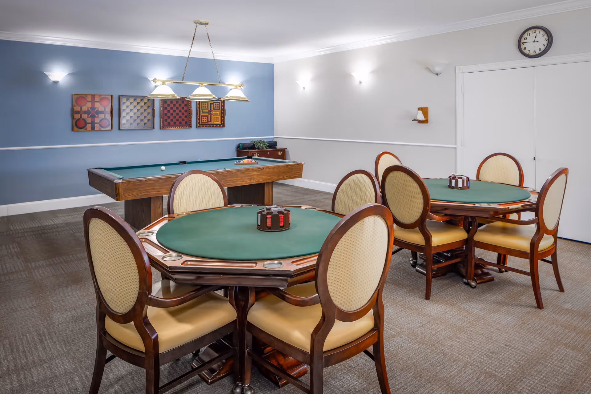 A game room with two round poker tables each surrounded by four beige cushioned chairs with wooden frames. Behind the tables is a pool table with a green felt surface. The walls are painted blue and white with four framed artworks hanging on the blue wall. A clock is mounted on the white wall above a closed door. The room is carpeted and well-lit with ceiling lights and wall sconces.