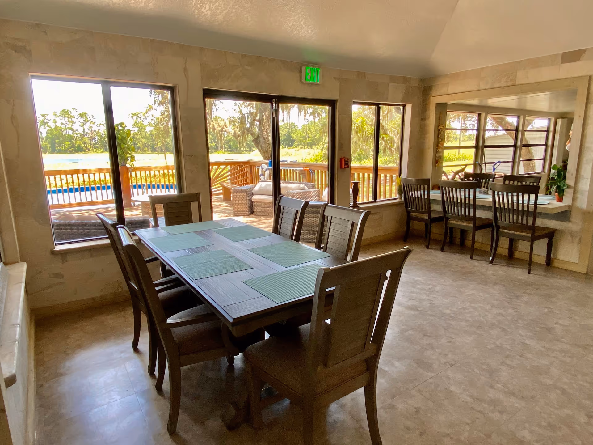 A bright dining room with a wooden table set with six chairs and green placemats. Large windows and a glass door provide a view of an outdoor deck with wicker furniture and a scenic landscape with trees and a body of water. There is also a counter with four chairs along a large interior window.