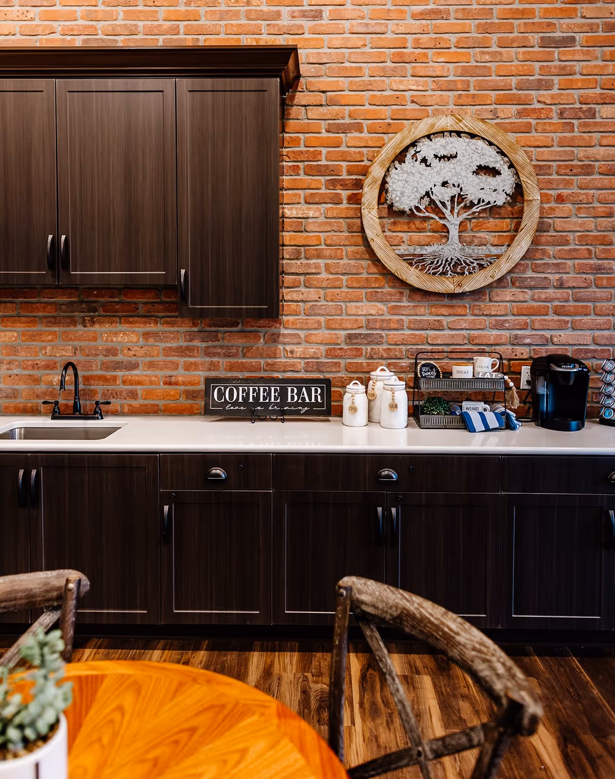 A cozy coffee bar setup with dark wooden cabinets, a white countertop, and a brick wall background. The countertop features a sink, a coffee maker, jars labeled coffee, tea, and sugar, and a small rack with coffee supplies. A decorative wooden and metal tree wall art hangs above the counter. In the foreground, part of a wooden table and rustic wooden chairs are visible.
