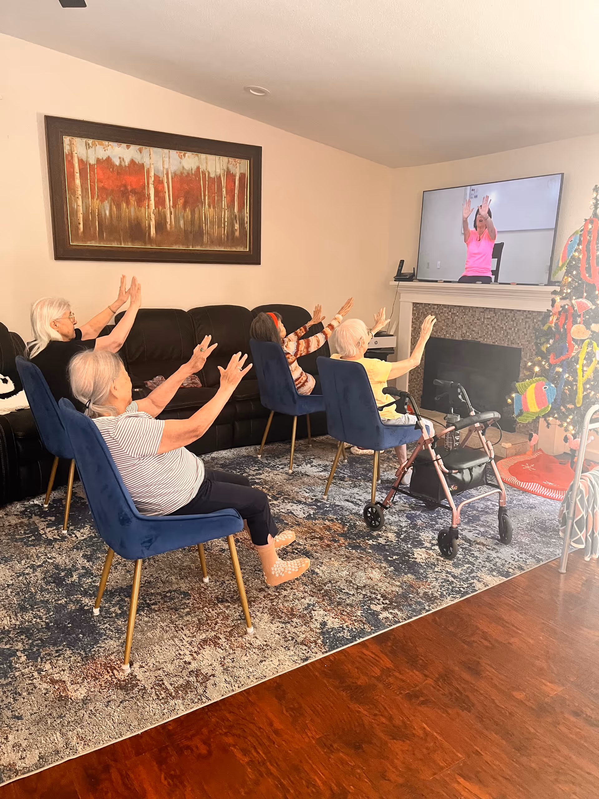 A group of elderly women seated in a living room on blue chairs and a black couch, participating in a seated exercise session by following a woman on a television screen. The room has a patterned rug, a fireplace, a decorated Christmas tree, and a large framed painting on the wall.