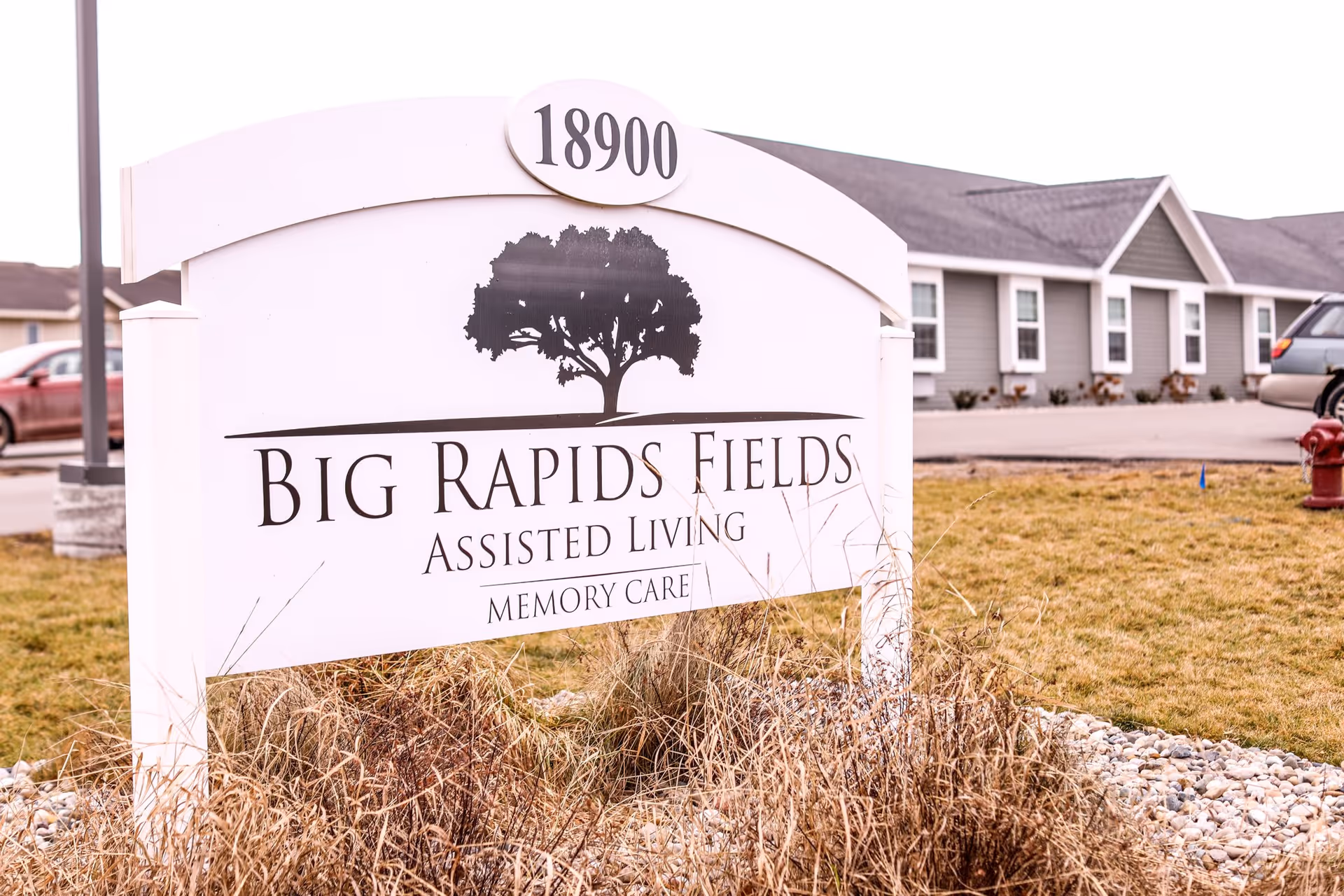 Outdoor view of a white sign with the address 18900 and the name Big Rapids Fields Assisted Living Memory Care, with a tree logo above the text. In the background, there is a gray building with white-trimmed windows, a parking area, and some parked cars. The foreground has dry grass and small rocks.