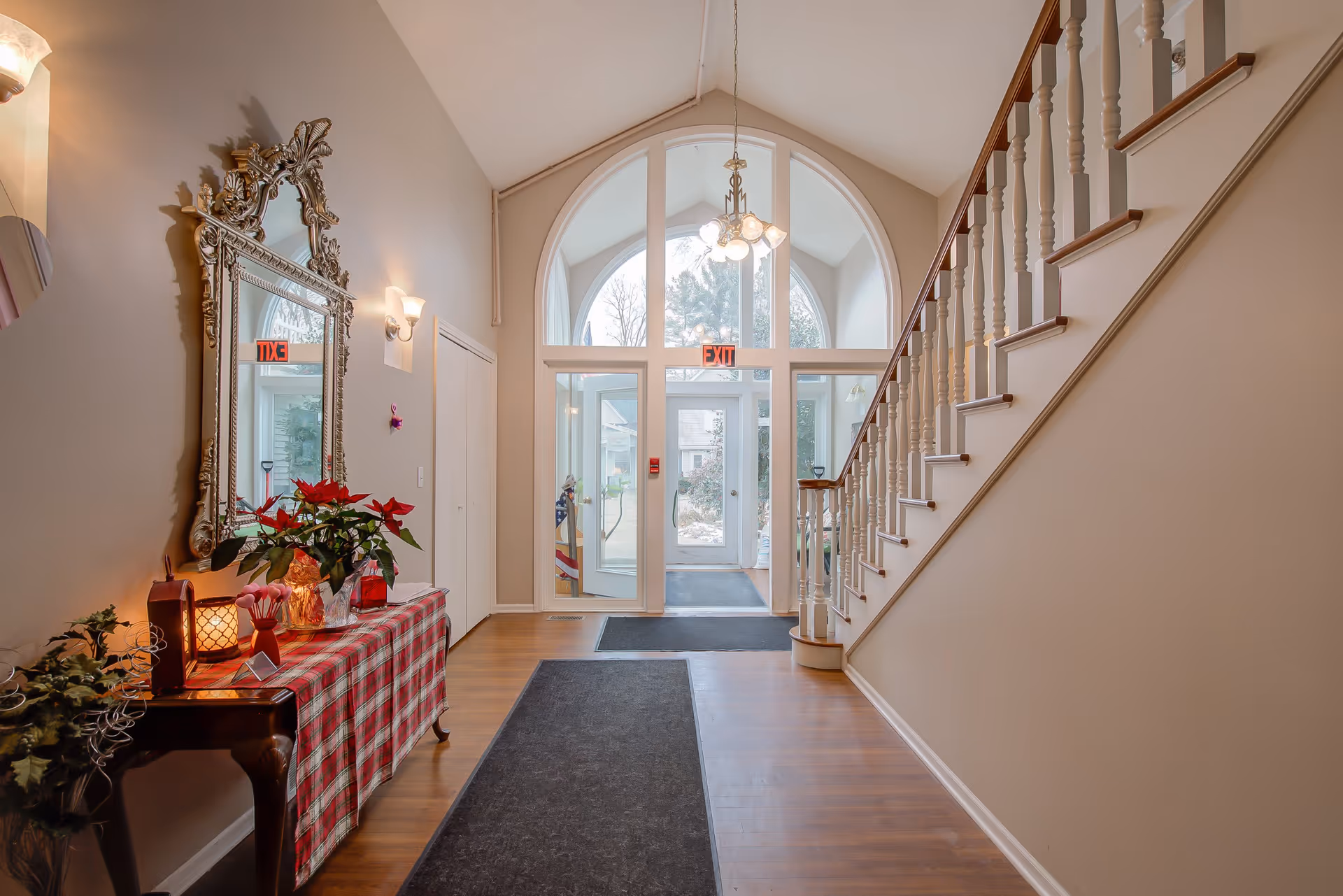 Interior view of an entrance hallway with a staircase on the right, a large decorative mirror above a table with a red plaid cloth and holiday decorations on the left, and a glass door with an exit sign at the far end.