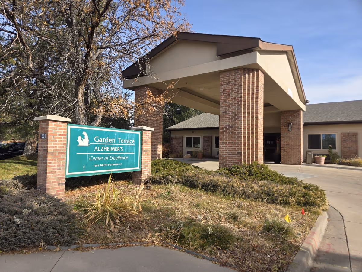 Front entrance of Garden Terrace Alzheimer's care center showing a green sign, brick columns supporting a covered driveway, and surrounding landscaping.