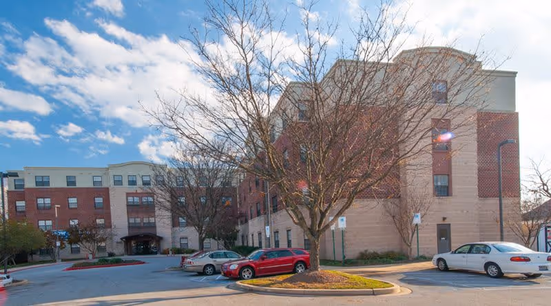 Exterior view of a multi-story senior living facility building with a parking lot in front. Several cars are parked, and leafless trees are visible under a partly cloudy blue sky.