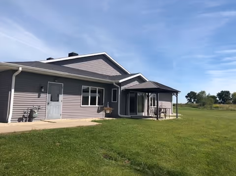 Single-story gray siding assisted living building with a covered patio and an expansive lawn under a clear blue sky.