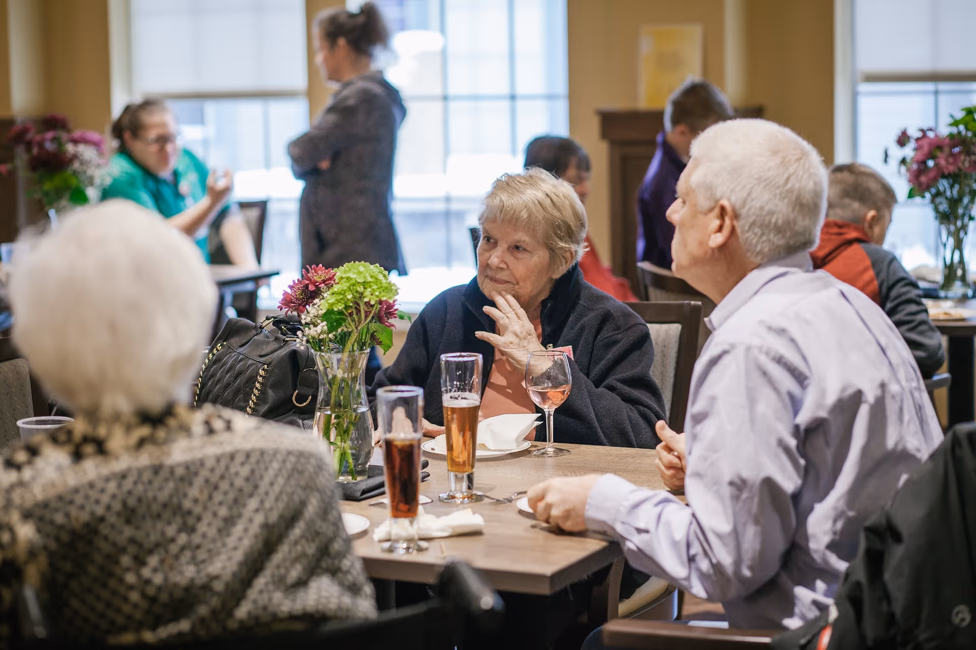 A group of elderly people sitting around a table in a dining area, engaged in conversation. The table has drinks and a vase with flowers. Other people are visible in the background, with large windows letting in natural light.