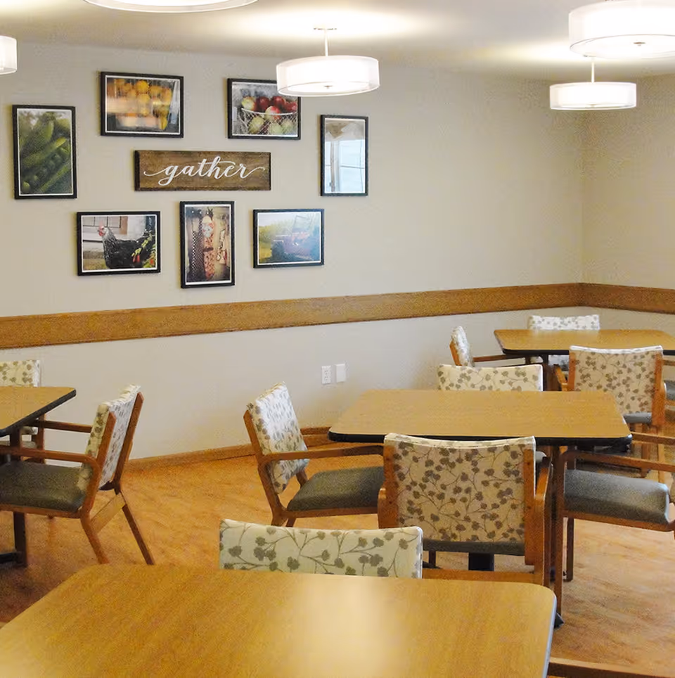 A dining room with several wooden tables and chairs that have floral patterned cushions. The walls are decorated with framed pictures of fruits, vegetables, and farm scenes, along with a wooden sign that says 'gather'. The room has light-colored walls, wooden flooring, and modern ceiling lights.