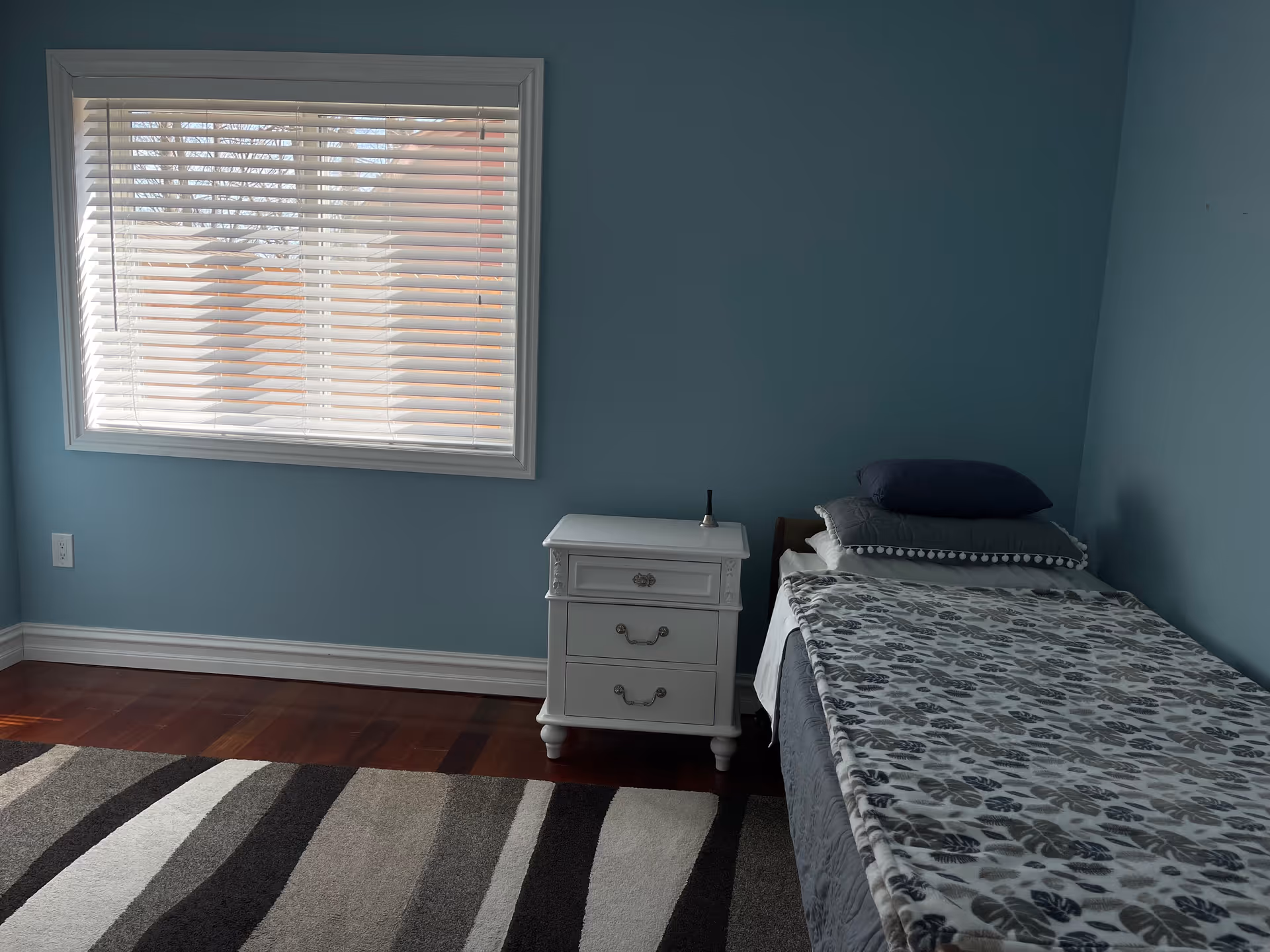 A simple bedroom with light blue walls, a window with white blinds, a single bed with patterned bedding and pillows, a white nightstand with drawers, and a striped area rug on a wooden floor.