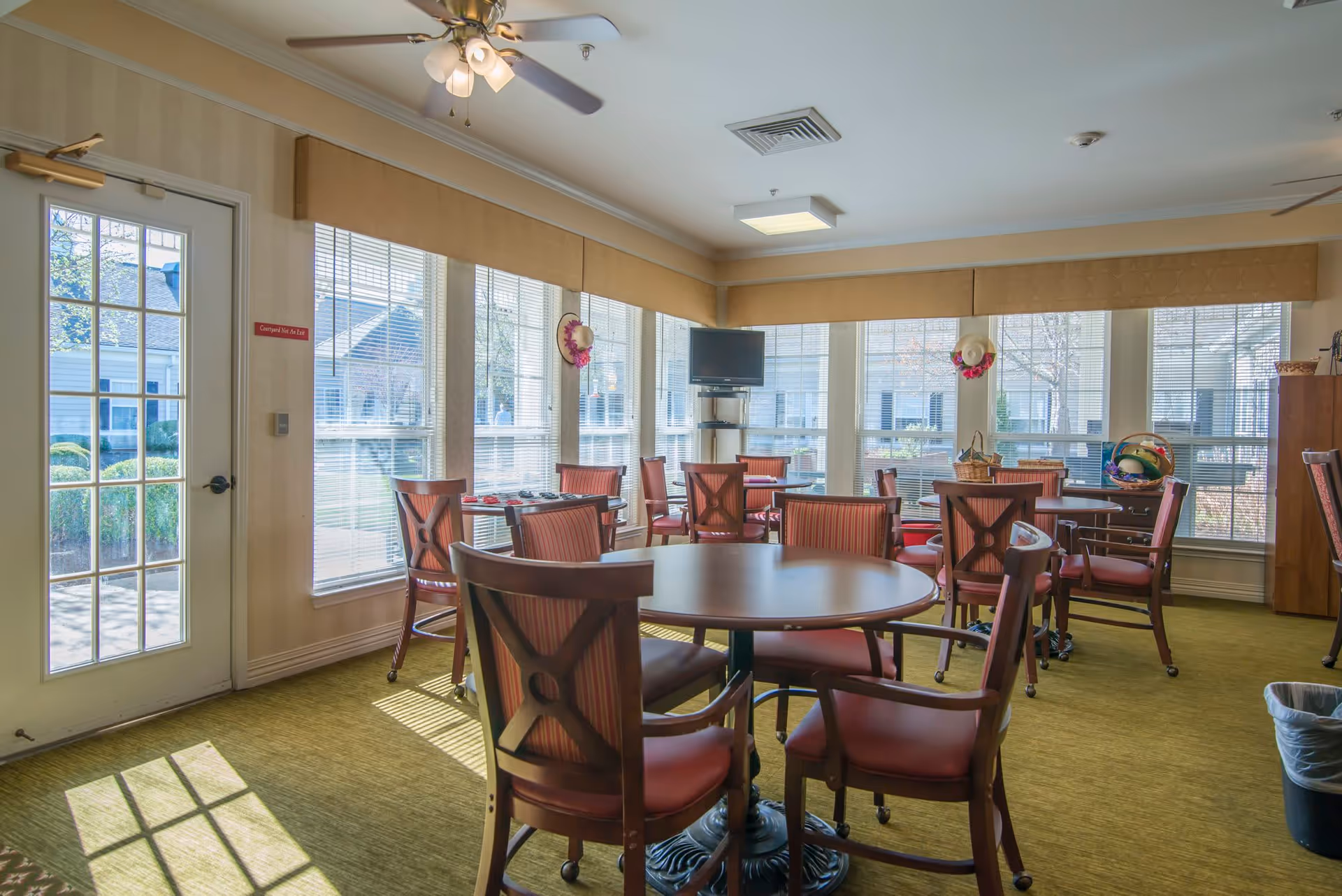 Bright dining/activity room with round tables, red-upholstered chairs, large windows, and a wall-mounted TV.